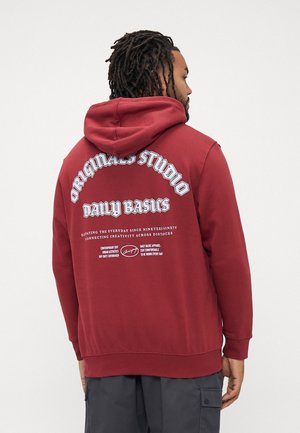 Mann mit Dreadlocks, der einen roten Kapuzenpullover mit dem Aufdruck "Originals Studio Daily Basics" auf dem Rücken trägt, kombiniert mit dunklen Cargo-Hosen.