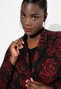 Černá bunda s červeným vzorem růží, texturovaná tkanina, zapínání na jeden knoflík a velký límec. Model drží klopu a má na sobě černý top.