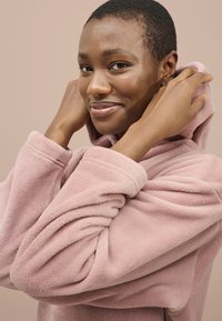 Lyserød fleece hoodie med en tekstureret overflade, oversized ærmer og en snoretrækshætte, båret af en person, der justerer stoffet.