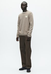 Lysebrun tekstureret sweater med 'W'-logo på brystet, kombineret med mørke olivengrønne bukser med vide ben og sorte sko, sat mod en hvid baggrund.