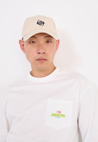 Jeune homme portant une casquette beige et une chemise à manches longues blanche avec un logo coloré "The North Face Expedition Series" sur la poche de la chemise.