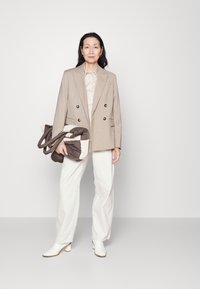 Beige dubbelknäppt kavaj, ljus skjorta, vita vida byxor och vita klackboots. Brun och krämfärgad totebag i handen.