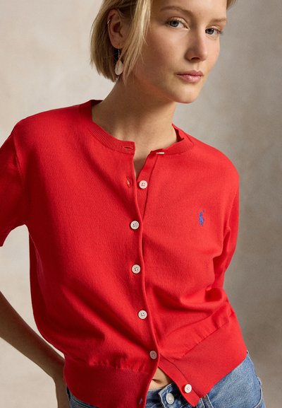 Nő piros gombos kardigánban, kék logóval és világoskék farmernadrágban, semleges háttér előtt állva.