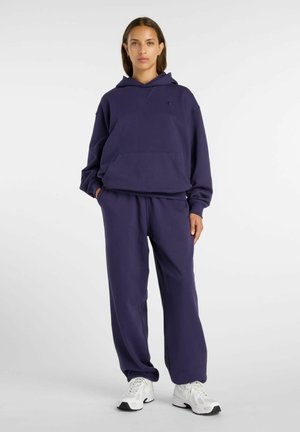 Vrouw staand, gekleed in een donkerblauwe hoodie en bijpassende joggingbroek met witte sneakers, tegen een effen lichte achtergrond.