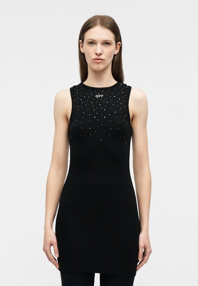 Γυναίκα που φοράει μαύρο αμάνικο φόρεμα με μικρές λευκές βούλες και το κείμενο «OFF» κοντά στη λαιμόκοψη, στέκεται μπροστά σε απλό ανοιχτόφωτο φόντο.