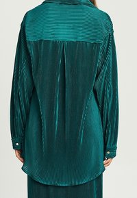 Camicia verde acqua a maniche lunghe, con pieghe verticali e polsini button-down, indossata dentro i pantaloni, vista di schiena su una persona in piedi.