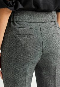 Gros plan sur une personne portant un pantalon taille haute à carreaux gris avec des poches passepoilées et un haut noir rentré.