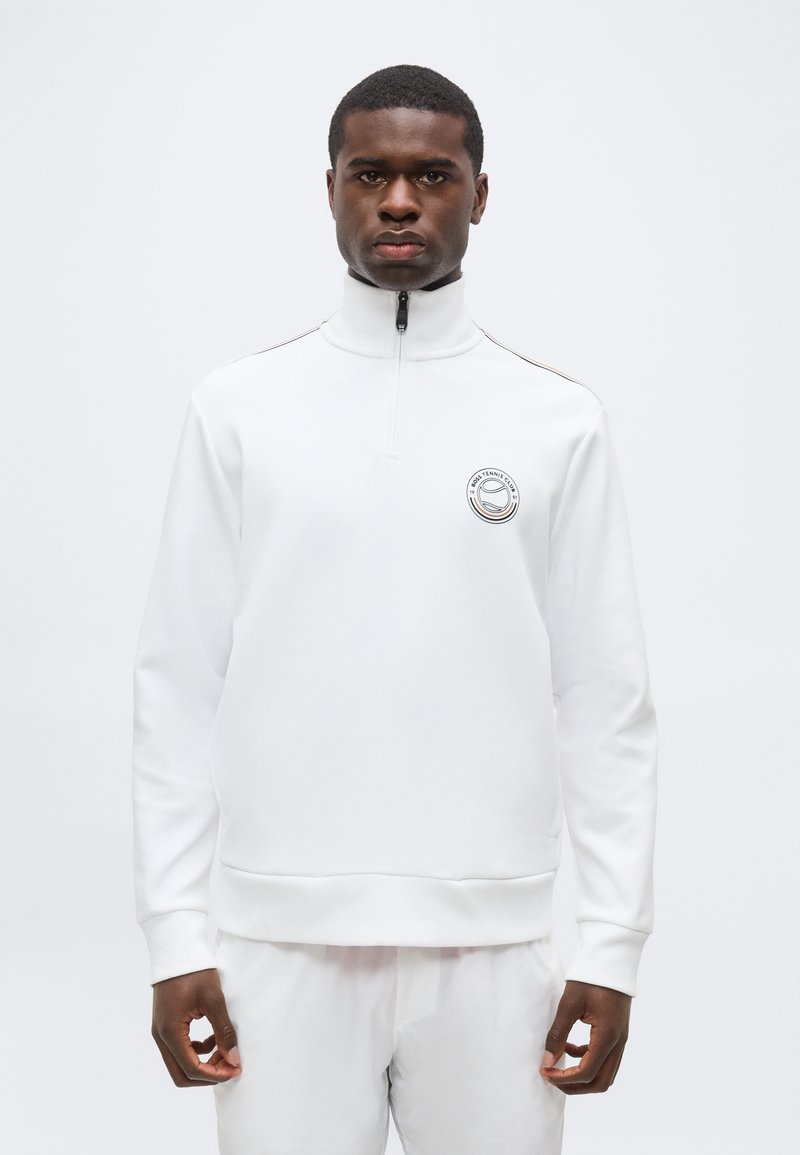 Homme portant un pull blanc à manches longues avec une fermeture éclair jusqu'à la poitrine et un logo de club de tennis sur la poitrine, debout devant un fond blanc uni.