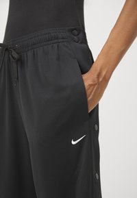 Personne portant un pantalon noir Nike avec taille élastique, boutons-pression latéraux et main placée dans la poche, sur un fond clair uni.