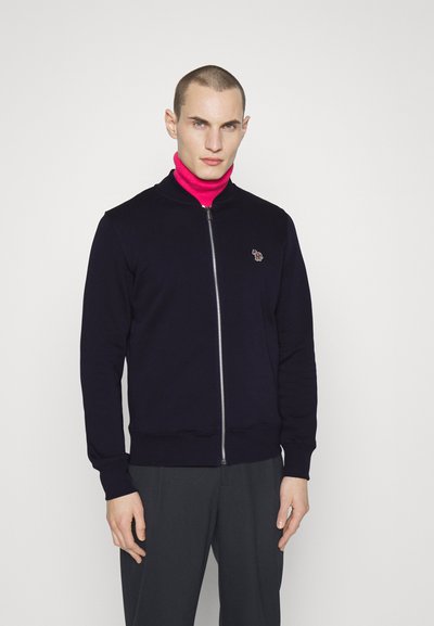 Marineblå zip-up sweatshirt med ståkrave, prydet med et diskret broderet design på brystet, båret over en lys pink turtleneck.