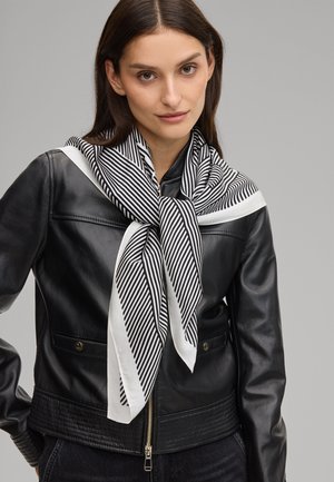 Woman wearing a black leather jacket and a black-and-white striped scarf tied around her neck against a plain gray background.