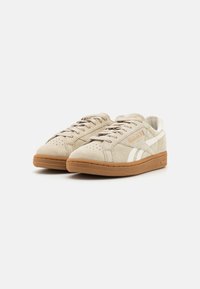 Beige mockask Reebok-sneakers med gummisulor och vita sidostreck, visade från framsidan på vit bakgrund.