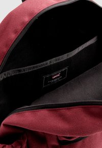 Εσωτερικό σακιδίου πλάτης από μπορντό ύφασμα με μαύρη επένδυση, που δείχνει μια ετικέτα με την επιγραφή "Levi's, San Francisco, Cal." και μια λεία άκρη φερμουάρ.