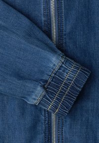 Ærme af en blå denimjakke med elastisk manchet og central metallynlås med gule syningsdetaljer.