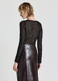 Top lungo in pizzo nero con maniche lunghe e motivo a diamante, abbinato a una gonna in vinile marrone lucido. Il tessuto è leggero e semi-trasparente.