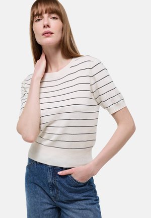 Femme portant un pull blanc à manches courtes avec de fines rayures horizontales noires et un jean bleu, posant avec une main sur le cou et l'autre dans la poche.
