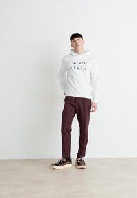 Bela bombažna kapuca z natisnjenim "CALVIN KLEIN" v črni barvi, v kombinaciji z temno burgundskimi hlačami in črnimi supergami z bež podplati ter roza vezalkami.
