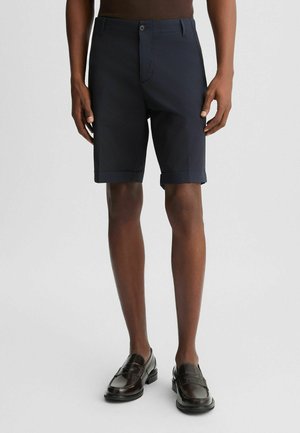 Pantaloni scurți din navy albastru, ajustați, cu front butonat, o pliă singulară și manșete rulate. Purtați cu loafers negri lustruiți, pe un fundal neutru.
