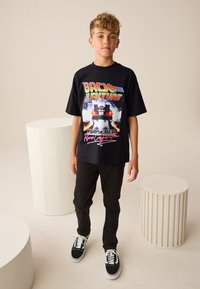 Tricou grafic negru cu design "Back to the Future" care prezintă o mașină și flăcări. Asortat cu pantaloni negri și adidași negri.