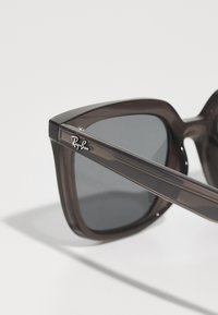 Ochelari Ray-Ban cu un cadru din plastic transparent de culoare maro închis, formă angulară supradimensionată, lentile întunecate și un accent cu logo argintiu pe braț.