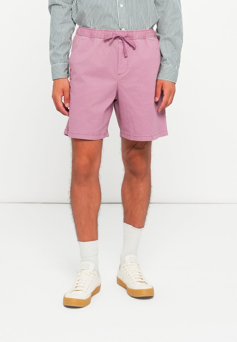 Lys rosa shorts med snor i taljen, glat overflade og opfoldet kant, parret med hvide sokker og beige sneakers.