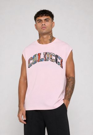 Hombre con cabello rizado y tatuajes, que lleva una camiseta sin mangas rosa con texto colorido "COLUCCI", de pie contra un fondo neutro.