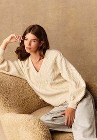 Krämfärgad stickad tröja med v-ringning, med texturerade mönster och små dekorativa detaljer. Modell som sitter i en beige tygfåtölj.