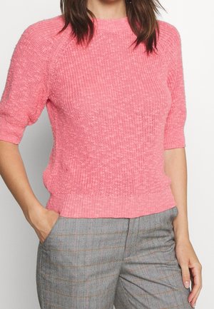 Vrouw die een roze gebreide trui met korte mouwen en grijze geruite broek draagt, met één hand in de zak, bijgesneden ter hoogte van de nek en halverwege het dijbeen.
