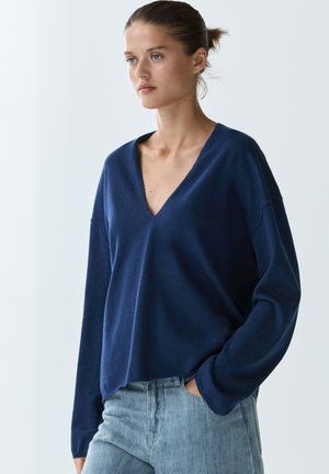 Frau mit zurückgebundenem Haar, die einen locker sitzenden navyblauen V-Ausschnitt-Pullover und hellblaue Jeans trägt, mit einer Hand in der Tasche vor einfachem Hintergrund.