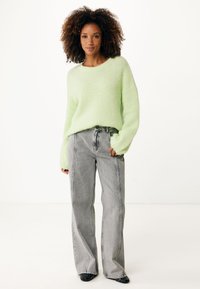 Světle zelený, chlupatý pletený svetr s širokými rukávy, v kombinaci s vysokými, volnými šedými džíny s kontrastními švy.