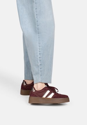 Personne portant un jean bleu clair et des baskets rouge foncé avec des rayures blanches et des semelles marron, debout sur un fond blanc.