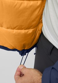 La veste rembourrée orange présente une texture lisse avec un ourlet bleu. Les cordons de serrage ajustables à la taille sont montrés en train d'être resserrés, améliorant ainsi l'ajustement.