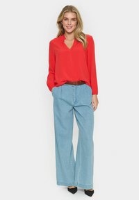 Blusa rossa a maniche lunghe con scollo a V, abbinata a pantaloni in denim a gamba larga di colore blu chiaro. Una cintura marrone e scarpe col tacco nere completano l'outfit.