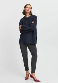 Pull en tricot marine avec un motif texturé, fentes sur les côtés, associé à un pantalon tailleur gris foncé et à des escarpins bordeaux.