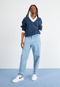 Casaco estilo corta-ventos azul-marinho com gola e punhos brancos. Jeans azul-claro de corte descontraído, combinados com ténis brancos com um swoosh azul-marinho.