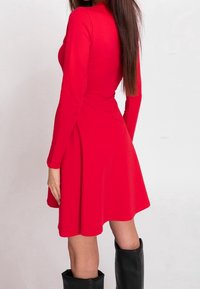 Rotes langärmeliges Kleid mit schwingendem Rock. Hergestellt aus glattem Stoff, hat es ein tailliertes Oberteil und einen runden Ausschnitt, getragen mit knielangen schwarzen Stiefeln.