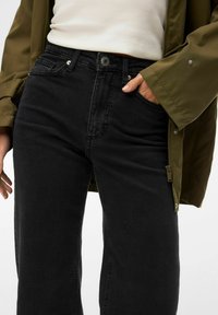 Personne portant un jean noir taille haute, un haut blanc côtelé et une veste vert olive, une main dans la poche, sur un fond uni.