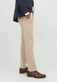Pantalones de vestir beige con corte slim, textura suave, con bolsillos laterales, combinados con zapatos de cuero marrón y un blazer azul marino parcialmente visible.