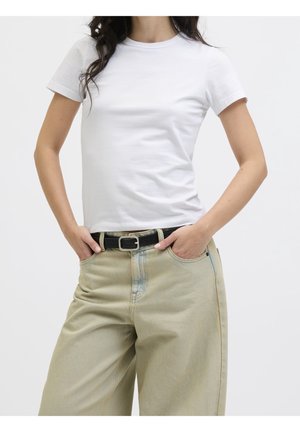 Mujer con camiseta blanca de manga corta metida en vaqueros beige de tiro alto con cinturón negro, manos en los bolsillos delanteros, fondo blanco.