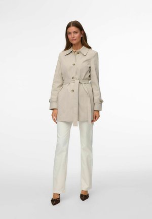 Žena v béžovom trenčkot s opaskom, biele nohavice a tmavé špicaté topánky, stojaca pred jednoduchým bielym pozadím.