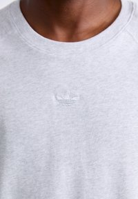 Tuvumā redzams cilvēks, kurš valkā gaiši pelēku krekla tipa sudrabu, uz krūtīm ir maza, diskrēta izšūta Adidas trefoil logo.