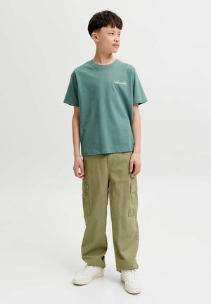 Nastolatek stoi, ubrany w turkusowy T-shirt, oliwkowe spodnie bojówki i białe trampki, patrzy w prawo na jednolitym tle.