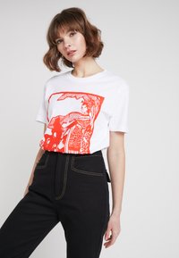 Vit t-shirt med en orange grafisk tryck med abstrakta figurer och mönster. Svarta höga byxor med kontrasterande sömmar.