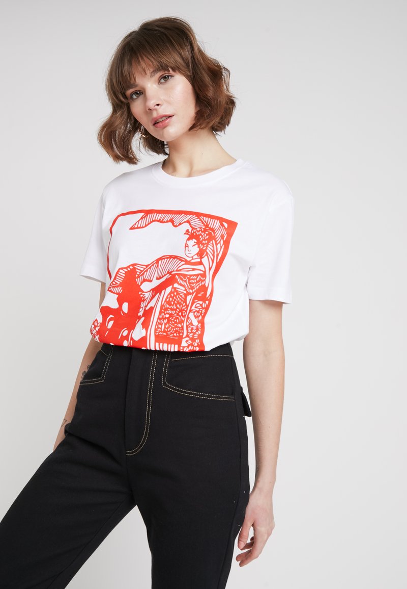 Vit t-shirt med en orange grafisk tryck med abstrakta figurer och mönster. Svarta höga byxor med kontrasterande sömmar.