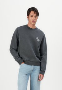 Mørkegrå sweatshirt med ribbet halsudskæring og manchetter. Har et lille broderet design på venstre bryst. Kombinationen er med lyseblå jeans.