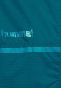 Close-up of teal fabric with stitched seam, horizontal reflective strip, and partially visible shiny "hummel" logo.