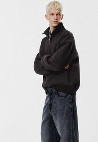Svart sweatshirt med hög krage och halvlång dragkedja, kombinerad med grå jeans med tunna ränder. Tyget verkar mjukt med en avslappnad passformsdesign.