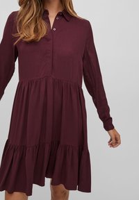 Abito bordeaux a maniche lunghe con colletto a bottoni, orlo a balze a strati, tessuto morbido e vestibilità rilassata, che mette in mostra una texture liscia e un design fluido.