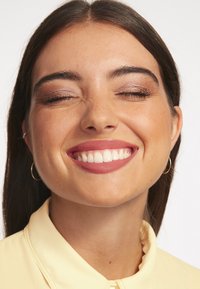 Närbild av en person som ler, med mellanbrunt hår, slät hy och lätt smink. De har små hoopörhängen och en ljusgul krageklädd topp.