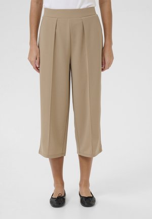Pantalon beige taille haute, raccourci et jambes larges, porté avec des ballerines noires sur une personne debout devant un fond blanc.